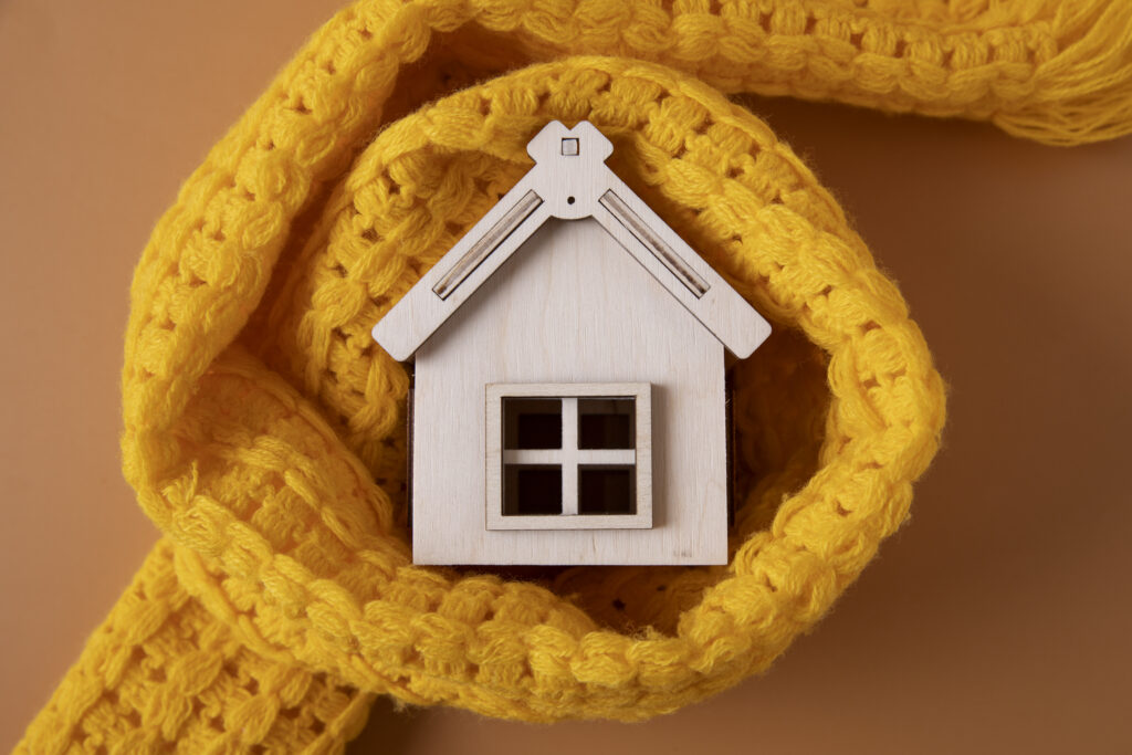 A small wooden toy house wrapped with a bright and cozy yellow knit scarf—symbolizing the warmth of a home heating system.