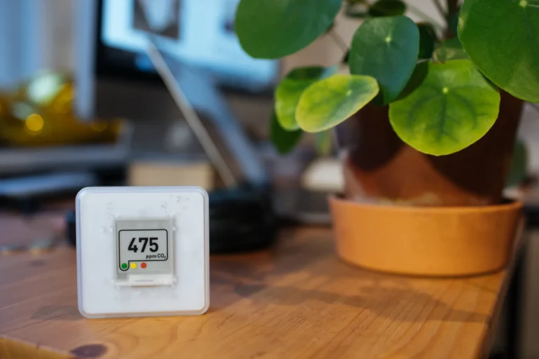 A digital CO2 monitor reading 475 ppm rests on a wooden desk beside a potted plant at Perfect Plumbing, Heating & Air's office.