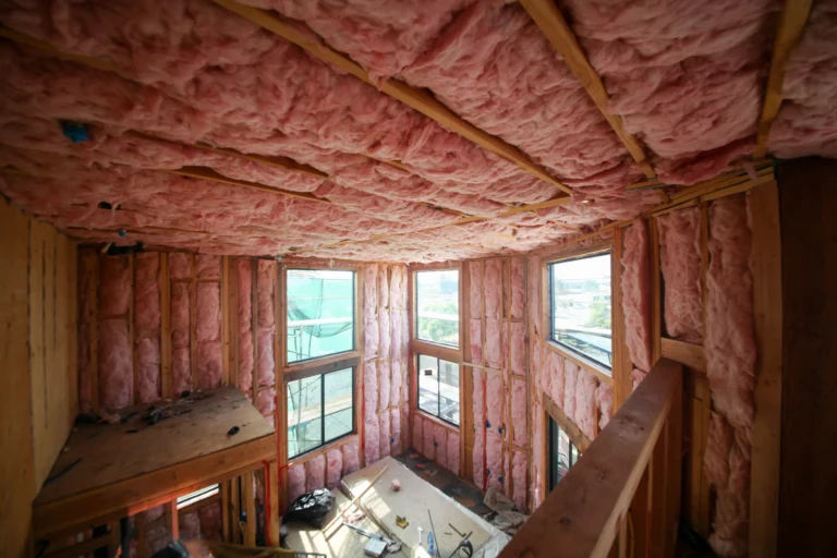 A bright and spacious home under construction showcases fresh pink fiberglass insulation along the walls and ceiling, with large windows letting in natural light. Construction materials neatly cover the floor, highlighting Perfect Plumbing, Heating & Air’s commitment to quality service throughout every step of your building project.